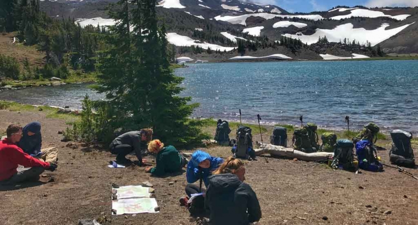 a group of backpacking students rest beside an alpine lake with snowy mountains in the background